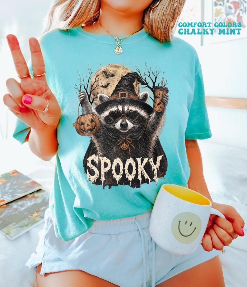 Pu&ograve; includere: Una t-shirt a maniche corte color verde menta gessato con la grafica di un procione che indossa un cappello da strega e un mantello, mentre tiene in mano un secchiello a forma di zucca e una bevanda. Include luna piena, alberi e la scritta "SPOOKY".