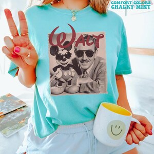 Op de afbeelding: Een mintgroen T-shirt met korte mouwen en een grafische print van Walt Disney en Mickey Mouse, beiden met een zonnebril op. Het woord Walt staat in een groot kastanjebruin lettertype boven de figuren. Het shirt heeft een ronde hals en een losse pasvorm.