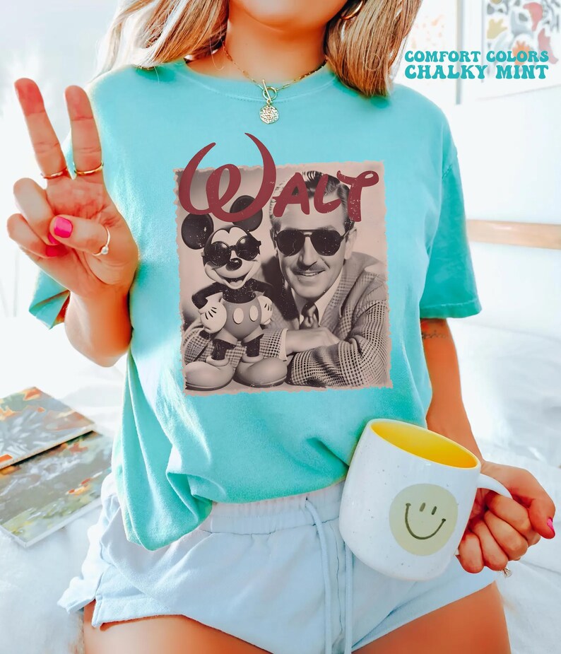 Op de afbeelding: Een mintgroen T-shirt met korte mouwen en een grafische print van Walt Disney en Mickey Mouse, beiden met een zonnebril op. Het woord Walt staat in een groot kastanjebruin lettertype boven de figuren. Het shirt heeft een ronde hals en een losse pasvorm.