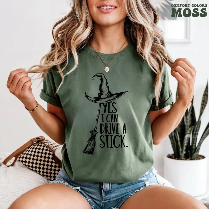 Pu&ograve; includere: T-shirt verde muschio con cappello da strega nero e grafica scopa. Il testo "YES I CAN DRIVE A STICK" &egrave; stampato sotto. La maglietta &egrave; un colore comfort.
