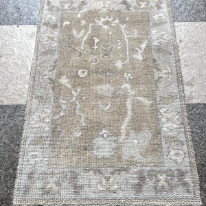 Puede incluir: Una alfombra rectangular beige con un diseño descolorido. La alfombra presenta un campo central con formas abstractas y un borde con motivos florales. Los bordes de la alfombra están acabados con flecos.