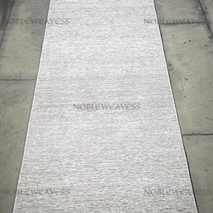 Puede incluir: Una alfombra larga y rectangular de color gris claro con una superficie texturizada. La alfombra tiene un sutil patrón de rayas que recorre la longitud. Las palabras "NOBLEWEAVESS" son visibles en la imagen. Adecuada para pasillos o entradas.