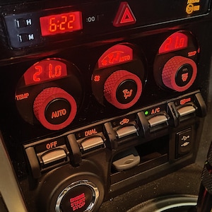 Könnte beinhalten: Ein schwarzes Armaturenbrett mit rot beleuchteten Knöpfen und einer Digitaluhr, die 6:22 anzeigt. Das Armaturenbrett verfügt über eine Temperaturanzeige, einen Auto-Knopf, einen Dual-Knopf, einen A/C-Knopf und einen roten Not-Stopp-Knopf.