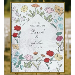 Op de afbeelding: Een ingelijste trouwbord op een gouden ezel. Het bord toont de tekst "THE WEDDING OF Sarah & John 10.18.26" in een huisvormige rand, omringd door kleurrijke bloemenillustraties.