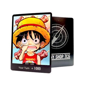 May include: A trading card depicting a cartoon character in a straw hat and red top, with the text "TAMAGO DON!! CARD" and "Your Turn + 1000". A second black card with a compass design is also visible.