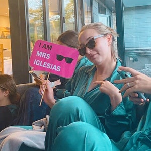 Op de afbeelding: Een vrouw in een tealblauwe satijnen ochtendjas en zonnebril houdt een roze bord vast met de tekst "I am Mrs. Iglesias" met een zonnebril erop.