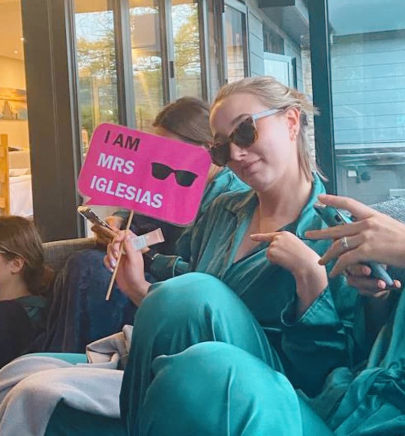 Op de afbeelding: Een vrouw in een tealblauwe satijnen ochtendjas en zonnebril houdt een roze bord vast met de tekst "I am Mrs. Iglesias" met een zonnebril erop.
