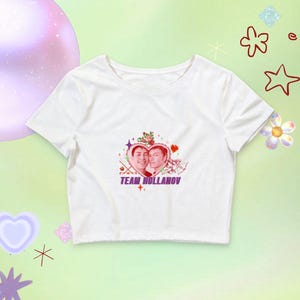 May include: A white short-sleeved crop top featuring a central graphic of two people inside a heart shape, surrounded by roses, stars, and Cupid illustrations. The text "TEAM HOLLANOV" is printed in purple below the heart graphic on the shirt.