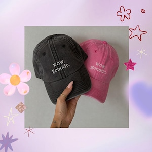 May include: Two baseball caps, one black and one pink, are displayed. Both caps have the phrase "wow. genetic." embroidered in white. The black cap is held in a hand. The caps are against a light background with decorative elements.