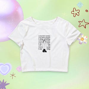 Könnte beinhalten: Weißes Crop-Top mit dem Text "I was born pretty. I've been pretty. I stay pretty." in Schwarz. Das Shirt zeigt eine Grafik von Frauenbeinen und High Heels. Der Hintergrund ist pastellgrün mit dekorativen Formen.