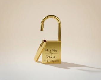 May include: A gold-colored padlock with an open shackle and a detached key. The lock is engraved with "Mr & Mrs Davis" and "est January 2022". The background is a solid, light beige color. The lock is a symbol of commitment and love.