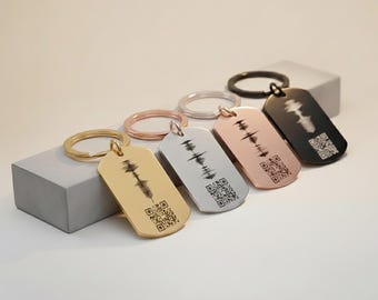 May include: A collection of personalized dog tag keychains in gold, silver, rose gold, and black. Each tag features a unique sound wave design and a QR code. The keychains are displayed on a light gray surface.