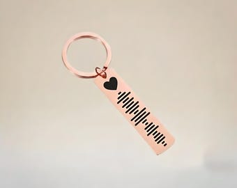 May include: A rose gold keychain with a black heart and a sound wave design. The keychain has a circular ring and a rectangular tag. The sound wave design is made of black vertical lines.