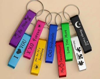 May include: A collection of colorful metal keychains with various engraved messages, including "I YOU", "Be Mine", and "Always Yourss". Each keychain has a silver ring and a rectangular bar in colors like purple, blue, and green.