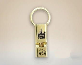 May include: Gold-colored Christmas-themed keychain. The rectangular pendant features a castle silhouette with the words "Merry Christmas" and a lower section with penguin and snowflake designs. The keychain has a circular ring.