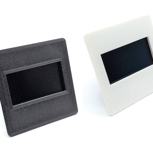 Puede incluir: Dos monturas para diapositivas, una negra y otra blanca, con una abertura rectangular para una diapositiva.