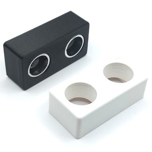 Puede incluir: Dos pequeños altavoces negros y blancos con aberturas redondas. Los altavoces están hechos de plástico y tienen una superficie texturizada.