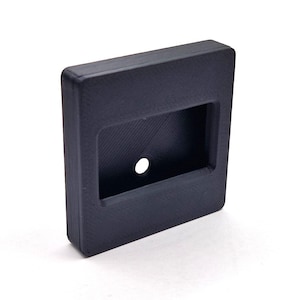 Może przedstawiać: Czarny, prostokątny przedmiot z wklęsłym prostokątnym obszarem i centralnym otworem. Obiekt ma teksturowaną powierzchnię i wydaje się być wykonany z litego materiału. Projekt jest prosty i funkcjonalny.