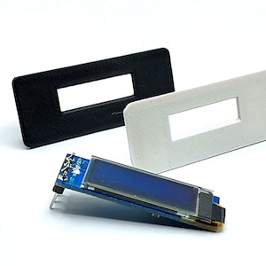 Peut inclure: Deux cadres en plastique imprimés en 3D, l'un noir et l'autre blanc, avec une découpe rectangulaire au centre. Un petit écran LCD bleu avec une carte de circuit imprimé est couché sur le côté devant les cadres.