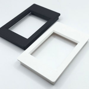 May include: Two rectangular frames, one black and one white, are positioned diagonally. Each frame has a recessed rectangular opening in the center. The frames are made of a matte material and sit on a white surface.