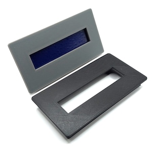 Op de afbeelding: Twee plastic frames met een rechthoekige uitsparing. Een frame is grijs en de andere is zwart.