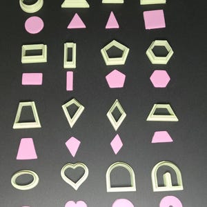 May include: A selection of light green plastic cookie cutters in diverse shapes, such as circles, triangles, squares, and hearts. Each cutter is accompanied by a pink shape of the same design, creating a contrasting colour scheme. The cutters are arranged on a dark surface.