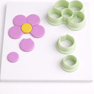 May include: A flower-shaped arrangement of purple fondant petals with a yellow centre, alongside green plastic flower-shaped cookie cutters. Additional purple fondant pieces and cookie cutters are also present.