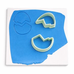 May include: A blue sheet of clay with three egg-shaped cookie cutters. One cutter is a full egg shape with a crack, and the other two are eggshell halves. The cutters are light green and sit on a white surface.