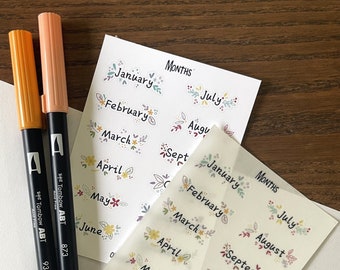 May include: Two sheets of stickers with floral designs and the months of the year written on them. The stickers are on a wooden surface with two black markers.