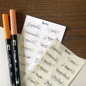 May include: Two sheets of stickers with floral designs and the months of the year written on them. The stickers are on a wooden surface with two black markers.