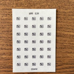 Könnte beinhalten: Eine Blatt mit transparenten Aufklebern mit einem schwarzen Umriss einer Kamera mit Blitz. Das Blatt hat den Text "MINI ICON" und "opigmom" aufgedruckt.