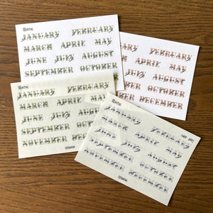 May include: Three sheets of stickers with the months of the year written in cursive script. The stickers are in three different color schemes: green and black, brown and black, and white and black.