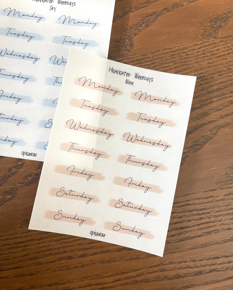 May include: Two sheets of stickers with the days of the week written in cursive. The top sheet has the days of the week in blue and white, while the bottom sheet has the days of the week in peach and white.