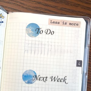 May include: A white gridded notebook with a blue and white sticker that says "To Do" and another sticker that says "Next Week". The notebook also has a sticker that says "Less is more".