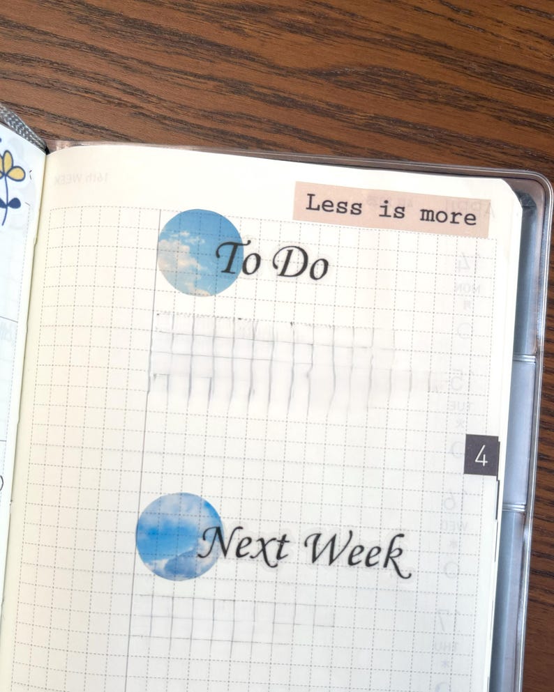 May include: A white gridded notebook with a blue and white sticker that says "To Do" and another sticker that says "Next Week". The notebook also has a sticker that says "Less is more".
