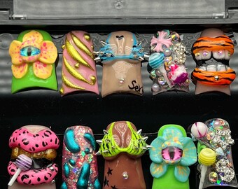 Puede incluir: Una colección de diez uñas artificiales con varios diseños. Las uñas presentan colores vibrantes, incluyendo rosa, verde y naranja, e incorporan elementos como púas, piedras preciosas y acentos de piruletas. Algunas uñas tienen el texto "Sex".