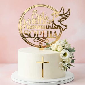 May include: A white frosted cake with a gold cross and a gold cake topper that reads "Erste Heilige Kommunion SOPHIA" with a dove design. The cake is decorated with white roses and greenery.