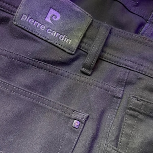 Pode incluir: Calças de ganga pretas com um pequeno remendo de couro preto que diz "pierre cardin" e um pequeno logotipo prateado com um design em espiral.