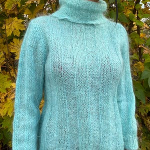 Puede incluir: Un suéter azul claro de manga larga con cuello alto. El suéter está hecho de un material suave y difuso. El fondo presenta hojas amarillas y verdes.