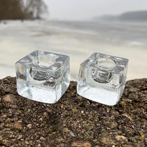 Könnte beinhalten: Zwei quadratische Kerzenhalter aus klarem Glas mit abgerundeten Kanten. Die Kerzenhalter stehen auf einer felsigen Oberfläche.