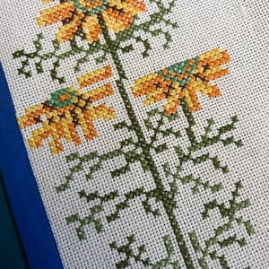Può includere: Primo piano di un ricamo a punto croce con un motivo floreale stilizzato. I fiori sono gialli, arancioni e verdi, e gli steli sono verdi. Il ricamo è su un tessuto bianco.