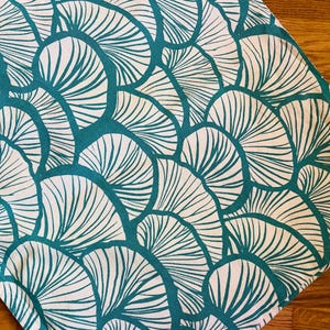 May include: A white and teal patterned textile featuring a repeating fan-like design. The teal outlines the fan shapes, creating a bold contrast against the white background. The fabric is displayed on a wooden surface.
