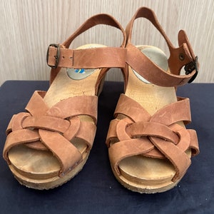 Könnte beinhalten: Ein Paar braune Ledersandalen mit einem geflochtenen Design an den Riemen. Die Sandalen haben eine Holzplattform und einen Schnallenverschluss.