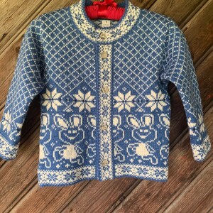 Op de afbeelding: Een blauwe en witte gebreide cardigan met een sneeuwvlok en geometrisch patroon. De cardigan heeft een knoopsluiting en lange mouwen.