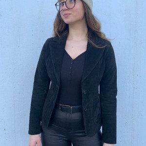 Può includere: Una donna indossa un blazer di velluto a coste nero con una maglietta nera e pantaloni neri. Indossa un berretto beige chiaro e occhiali.