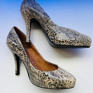 Pode incluir: Um par de sapatos de salto alto com padrão de pele de cobra em tons de castanho, bege e preto. Os saltos são finos e pretos, e os sapatos têm um design de bico arredondado. O interior do sapato é de cor castanho claro.