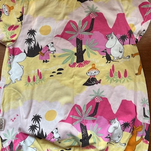 May include: Yellow short-sleeved shirt with a colorful landscape print featuring cartoon characters, pink mountains, palm trees, and a sun. The shirt has a round neckline and a fitted design, suitable for children's wear.