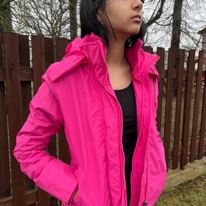 Könnte beinhalten: Eine leuchtend pinkfarbene Kapuzenjacke mit Reißverschluss. Die Jacke hat zwei Taschen vorne und einen Kordelzug in der Taille.