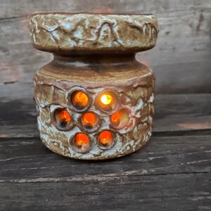 Puede incluir: Un portavelas de cerámica con un exterior texturizado marrón y beige. El portavelas tiene varias aberturas circulares que emiten un brillo naranja cálido de las velas encendidas en su interior. El diseño es rústico y decorativo.