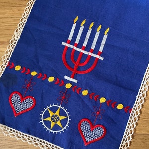 Puede incluir: Camino de mesa azul con bordados de menorá, corazones y estrellas. La menorá es roja con velas blancas y llamas amarillas. El camino tiene un ribete de encaje blanco.